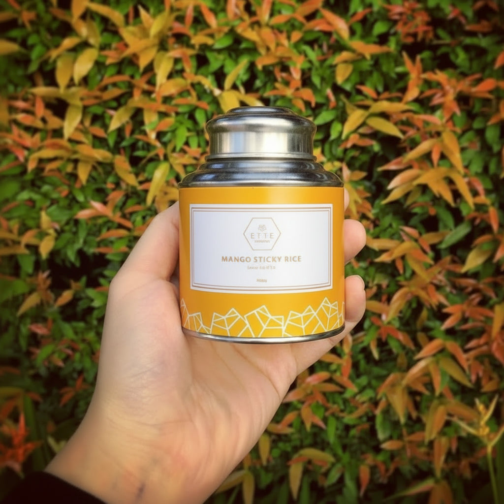 A hand holding a yellow tea can labeled 'Mango Sticky Rice' with a background of green and orange foliage.