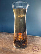Clear glass carafe with iced tea and floating tea leaves on a wooden surface with a blue background.