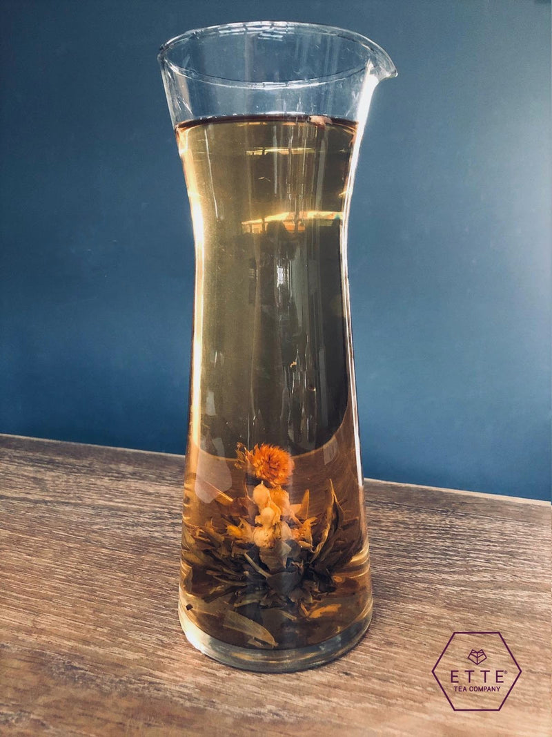 Clear glass carafe with iced tea and floating tea leaves on a wooden surface with a blue background.