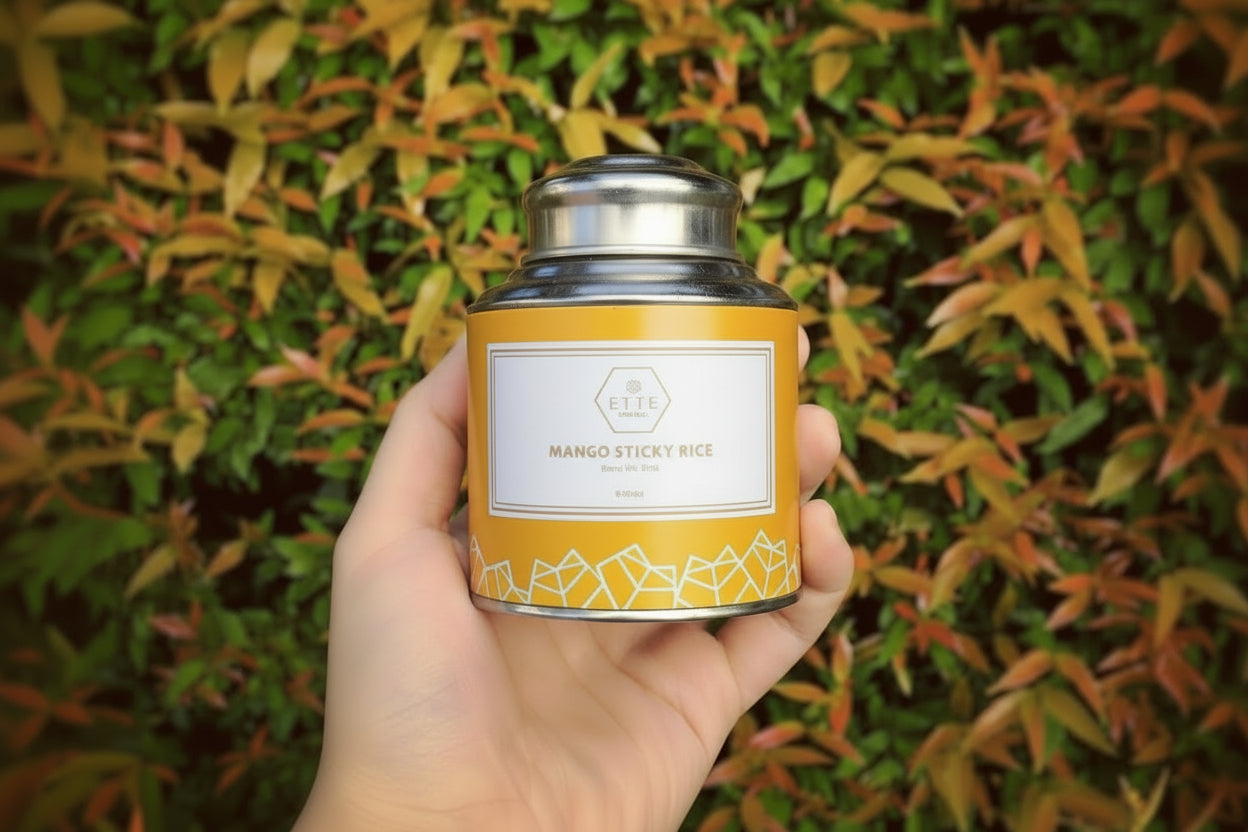 A hand holding a yellow tea can labeled 'Mango Sticky Rice' with a background of green and orange foliage.