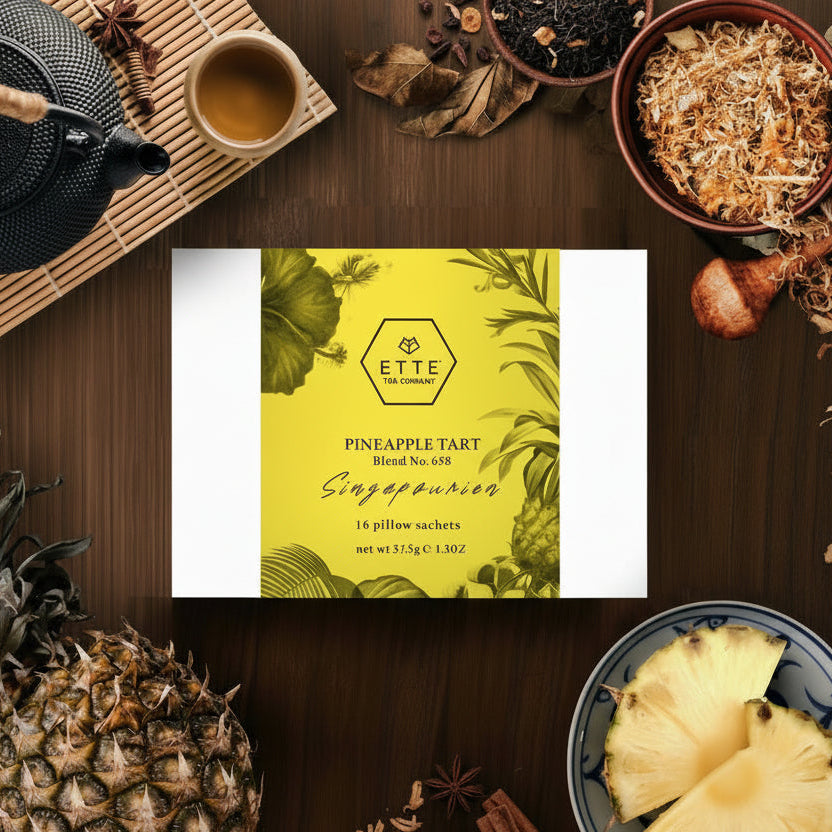 A box of ETTE Pineapple Tart flavored tea sachets, with visual elements suggesting a tropical theme, including pineapples and leaves. The box is presented on a table with tea making equipment and ingredients around it.