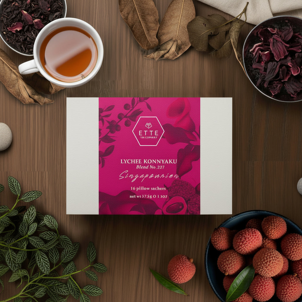 A box of Lychee Konnyaku tea sachets with a cup of brewed tea and various tea leaves and ingredients in the background, presented on a wooden table.